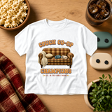 White T-shirt printed with a plaid retro couch holding popcorn and two game controllers, two stars and the words Couch Co-op Champions, Est. in the Family Room